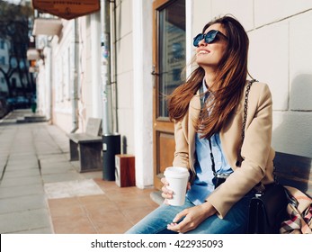 Beautiful and young girl in a coat and scarf and sunglasses sitting on the bench. Woman drinking coffee and laughing. Summer.