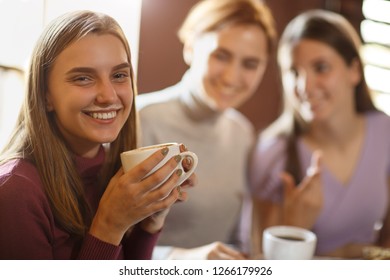 Beautiful Young Girl With Coffee Foam Over Her Upper Lip Smiling And Looking At Camera. Happy Woman Drinking Coffee From Large Cup, Holding In Hand, At Cafe With Two Girlfriends On Blurry Background.