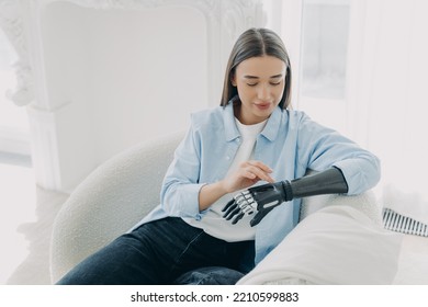 Beautiful young girl with disability setting her sensory bionic prosthetic arm, sitting in armchair. Modern woman using artificial robotic hand after limb loss.