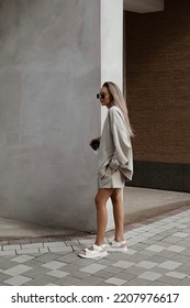 beautiful young girl dressed in beige knitted oversized suit with sweatshirt and shorts, sandals, bag, sunglasses, stylish fashion outfit, full length lifestyle model - Shutterstock ID 2207976617
