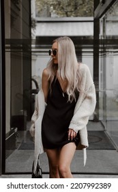 beautiful young girl dressed in black mini dress with straps, beige knitted oversized cardigan, massive boots, sunglasses, bag, accessories, wavy hair, stylish fashion outfit, lifestyle model