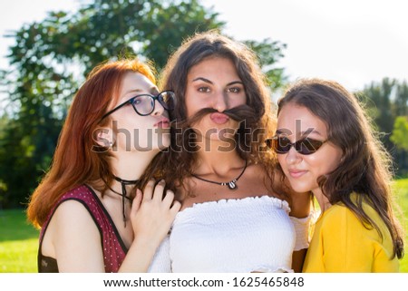 Beautiful young girl with her two friends, funny close-up front portrait in park, looking at camera and grimacing, holding her hair as a moustache.