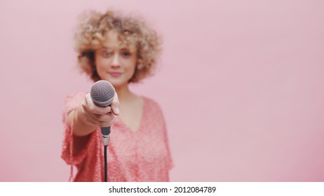 Beautiful young girl holding microphone towards the camera. The focus is on the mic while the girl is blurred in the background. Isolated with pink background studio. Reporter or Journalist concept.