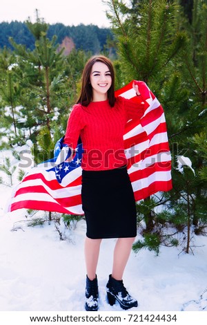 A beautiful young girl holds a flag of the United States of America. Winter in a snowy park with fir-trees. clothed in a chiffon knitted jacket.