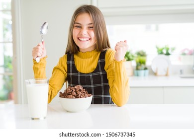 Beautiful young girl kid eating chocolate cereals and glass of milk for breakfast screaming proud and celebrating victory and success very excited, cheering emotion