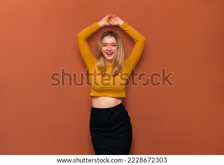 A beautiful young girl with large breasts, holds her hands above her head, laughs at the camera with her eyes narrowed, in a yellow blouse with bare belly and a black long skirt on an orange backdrop.