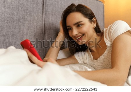 A beautiful young girl lies in bed and enjoys communication, holds a smartphone in her hands.