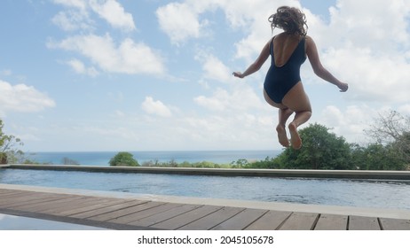 A Beautiful Young Girl With Long Hair And A Blue Swimsuit Jumps Into The Pool In Slow Motion, In An Expensive Villa With A Beautiful View Against The Background Of Blue Sky And White Clouds On A Day.