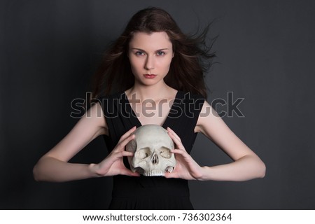 A beautiful, young girl with long hair in a black dress with a skull in her hands. Studio photo on a gray background. Witchcraft, necromancy, fortune telling, Halloween.