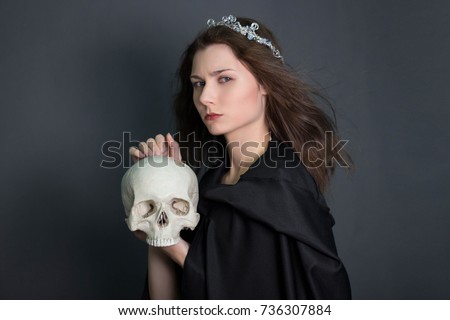 A beautiful, young girl with long hair in a black cloak with a hood and diadem on her head in her hands holds a skull. Studio photo on a gray background. Witchcraft, necromancy, divination, Halloween.
