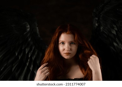 beautiful young girl with long red hair and black wings in a dark room - Shutterstock ID 2311774007