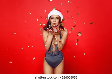 beautiful young girl model stands on red background in sexy bathing suit  and a Christmas Santa hat.On background flying golden confetti.