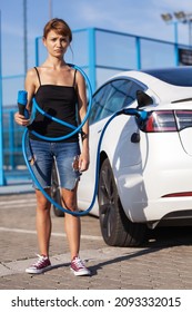 Beautiful Young Girl Next To An Electric Car. Clumsy Wrapped And Confused With A Charging Cable Around Her Body.