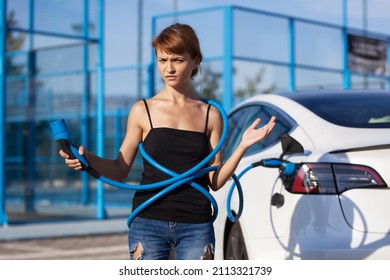 Beautiful Young Girl Next To An Electric Car. Clumsy Wrapped And Confused With A Charging Cable Around Her Body.