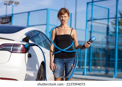 Beautiful Young Girl Next To An Electric Car. Clumsy Wrapped And Confused With A Charging Cable Around Her Body.