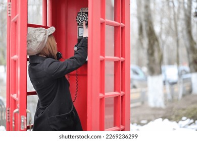 Beautiful young girl in a phone booth. The girl is talking on the phone from the payphone. English telephone booth in the street and a woman talking on the phone.