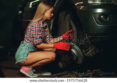 A beautiful young girl in a plaid shirt and short denim shorts is repairing a car at a service station. Modern independent girl. Car mechanic repairing a wheel.