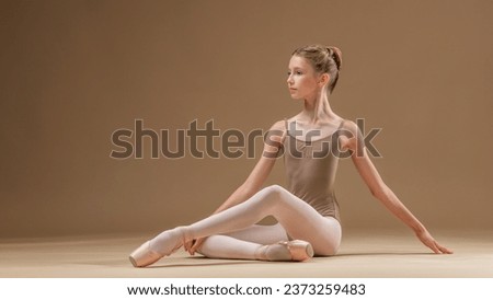 Beautiful young girl professional ballerina student in pointe shoes and leotard sitting posing on a light beige background.