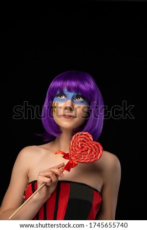 beautiful young girl in a purple wig, mask and a lollipop in her hands smiles and looks up on a black background. Event at a party, carnival or masquerade