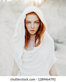 A beautiful young girl with red hair and freckles looks intently at the camera. Woman in the hood and clothes for the desert. Concept.