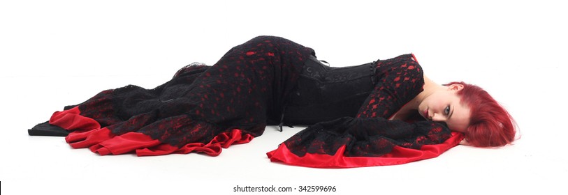 Beautiful young girl with red hair, wearing a long  gothic black and red flamenco lace gown with black corset. fallen or lying on the ground. isolated on white background. 