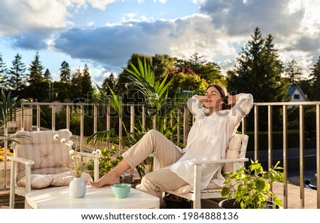 Beautiful young girl relaxing and enjoying sun while having tea, sitting at balcony at sunlight at summer. Slow living, tranquil moment, mental health. Backyard terrace vacation.