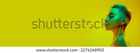 Beautiful young girl with short blonde hair posing with bare shoulders against yellow studio background in green neon light. Cyberpunk style. Banner. Concept of technology, modern life, digital art