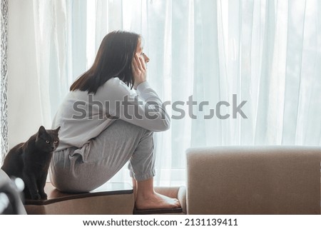 A beautiful young girl sits at home alone and is bored, she is sad, she looks out the window. Next to her is a gray cat with bright eyes. The concept of boredom, depression in young girls.