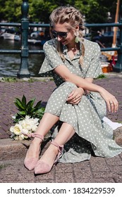 A beautiful young girl is sitting on the pavement in a fashion pose with a bouquet of white peony flowers in a long green dress and cute flat shoes with ties on the ankles. Street Style in Amsterdam