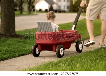 Beautiful young girl sitting in a red wagon cart by the road outdoors. Father is pulling a wagon with daugther. 