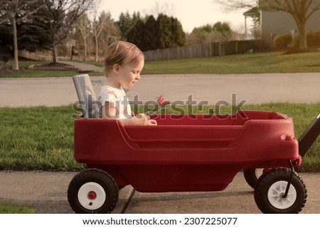 Beautiful young girl sitting in a red wagon cart by the road outdoors. Cute little girl  is holding a flower in her hand.
