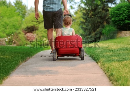 Beautiful young girl sitting in a red wagon cart outdoors. Father is pulling a red wagon with his daughter.