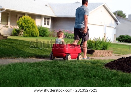 Beautiful young girl sitting in a red wagon cart outdoors. Father is pulling a red wagon with his daughter.