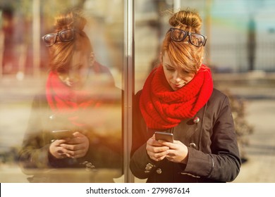 Beautiful young girl standing next to a shop window and typing a text message on her smart phone