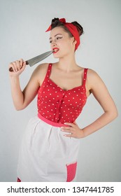 Beautiful Young Girl Standing In Red Polka Dot Pinup Dress And Apron, Holding A Big Knife And Picking Her Teeth Because The Food Is Stuck, On A White Studio Plain Background Alone