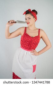 Beautiful Young Girl Standing In Red Polka Dot Pinup Dress And Apron, Holding A Big Knife And Picking Her Teeth Because The Food Is Stuck, On A White Studio Plain Background Alone