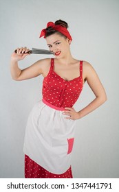 Beautiful Young Girl Standing In Red Polka Dot Pinup Dress And Apron, Holding A Big Knife And Picking Her Teeth Because The Food Is Stuck, On A White Studio Plain Background Alone