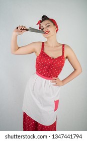 Beautiful Young Girl Standing In Red Polka Dot Pinup Dress And Apron, Holding A Big Knife And Picking Her Teeth Because The Food Is Stuck, On A White Studio Plain Background Alone