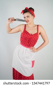 Beautiful Young Girl Standing In Red Polka Dot Pinup Dress And Apron, Holding A Big Knife And Picking Her Teeth Because The Food Is Stuck, On A White Studio Plain Background Alone