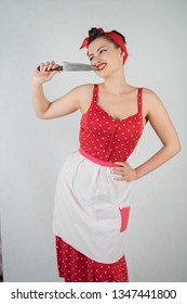 Beautiful Young Girl Standing In Red Polka Dot Pinup Dress And Apron, Holding A Big Knife And Picking Her Teeth Because The Food Is Stuck, On A White Studio Plain Background Alone