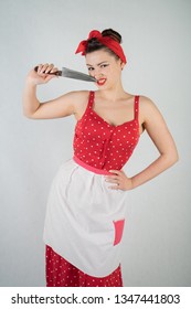 Beautiful Young Girl Standing In Red Polka Dot Pinup Dress And Apron, Holding A Big Knife And Picking Her Teeth Because The Food Is Stuck, On A White Studio Plain Background Alone