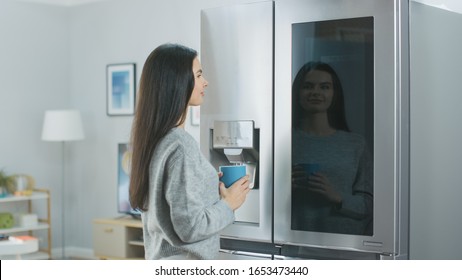 Beautiful Young Girl Stands Next to a Refrigerator While Drinking Her Morning Coffee. She is Checking the Weather Forecast and a To Do List on a Smart Fridge at Home. Kitchen is Bright and Cozy.
