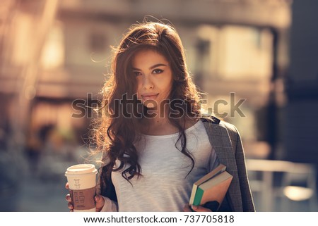Beautiful young girl student drinking caffee on the way to college or university. Fashion dressed for school carrying her books and notebook wearing sunglasses. Lifestyle in the city back to school