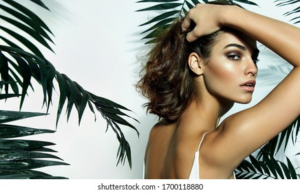 Beautiful young girl in the studio on a white background with dark skin, brown eyes and dark short hair holds a large green tropical leaf in her hands and covers part of her fac