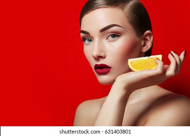 Beautiful young girl in the studio on an orange background holds in her hand a slice of orange. Smooth skin and hair. Styling, shiny hair. Fruits, health, diet, fashion, beauty. Makeup - red lips.