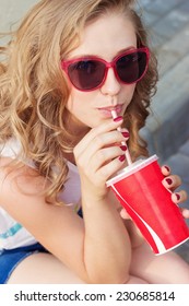 beautiful young girl in sunglasses sitting on the street and drink a coke with a paper Cup from the tube