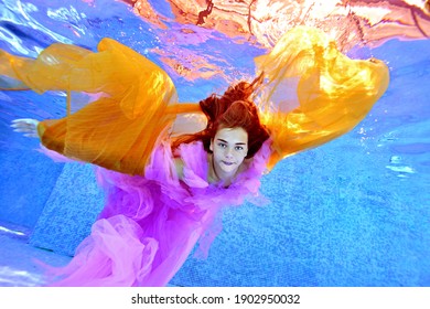 A Beautiful Young Girl Is Swimming Underwater In A Pool In A Purple Dress And With A Yellow Cloth In Her Hands. Her Arms Are Spread Out Like Wings. Underwater Fashion Photography.