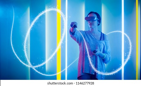 Beautiful Young Girl Wearing Virtual Reality Headset Draws Abstract Lines and Figures with Joysticks / Controllers. Creative Young Girl Does Concept Art with Augmented Reality. Neon Retro Lights