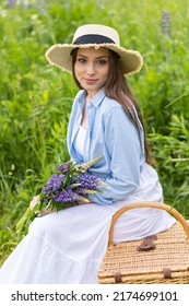 Beautiful Young Girl In A White Dress, Straw Hat With A Bouquet Of Violet Flowers In Her Hands And Picnic Basket. Pretty Woman In Summer In The Blooming Field Holding A Bunch Of Purple Lupin