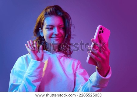 Beautiful young girl in white hoodie talking on video call with mobile phone against gradient blue purple studio background in neon light. Concept of emotions, youth, lifestyle, communication